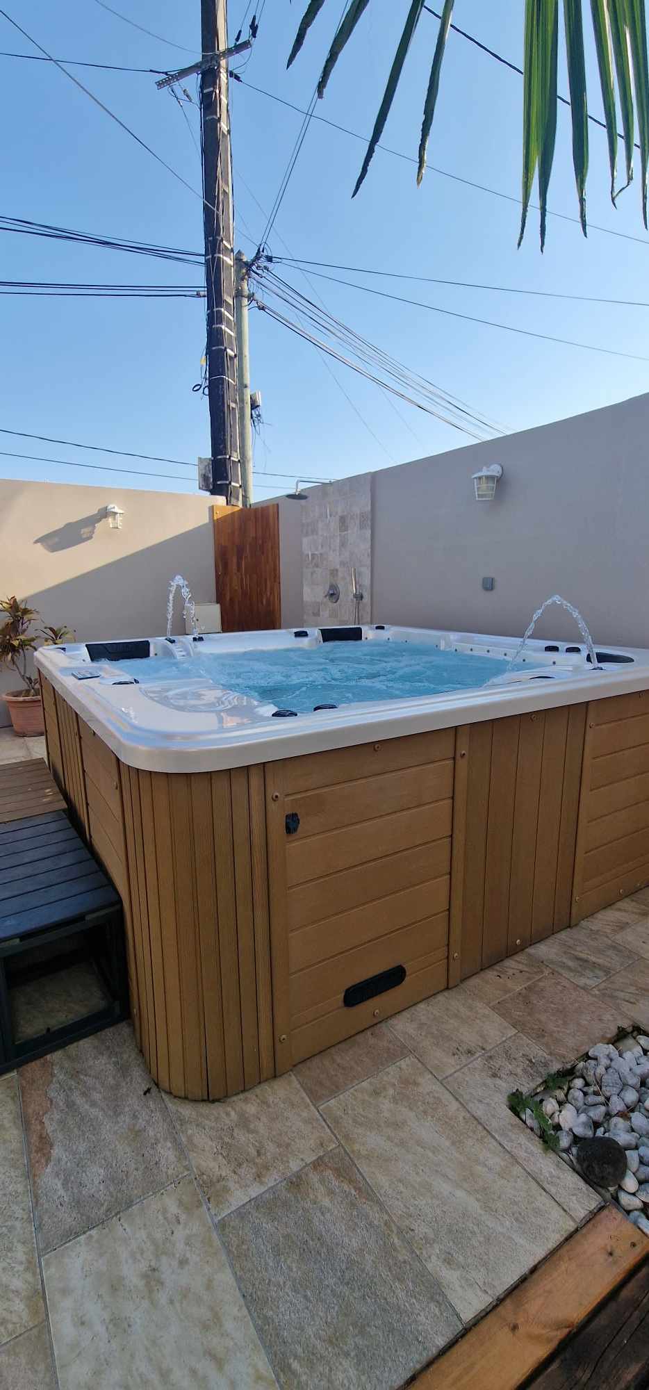 A spacious outdoor area features a jacuzzi with multiple jets, surrounded by natural stone tiles. Water jets create gentle splashes, enhancing the space. A wooden deck provides a comfortable area nearby, complemented by decorative white stones and soft light from a wall-mounted fixture.