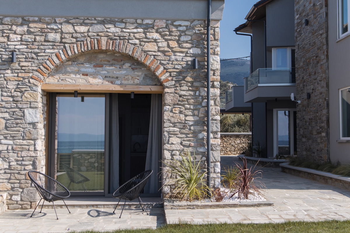 A stone exterior showcases a rustic architectural style, featuring an arched doorway with wooden accents. Two modern black chairs are positioned on a stone patio, offering scenic views of the surrounding landscape and the sea in the background.
