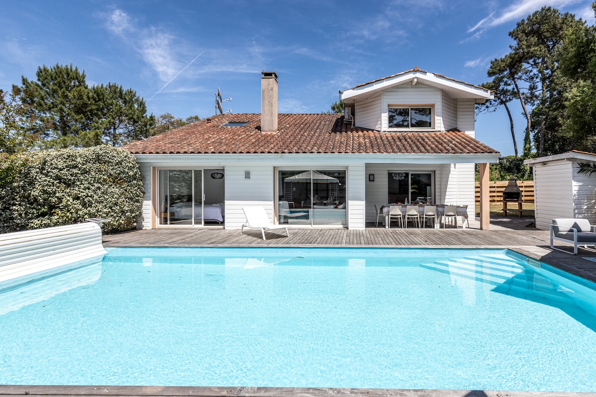 A spacious villa exterior is showcased, featuring a large swimming pool framed by a wooden deck. The house includes multiple glass doors allowing natural light to fill the interior. Outdoor dining furniture is visible, enhancing the inviting outdoor atmosphere.
