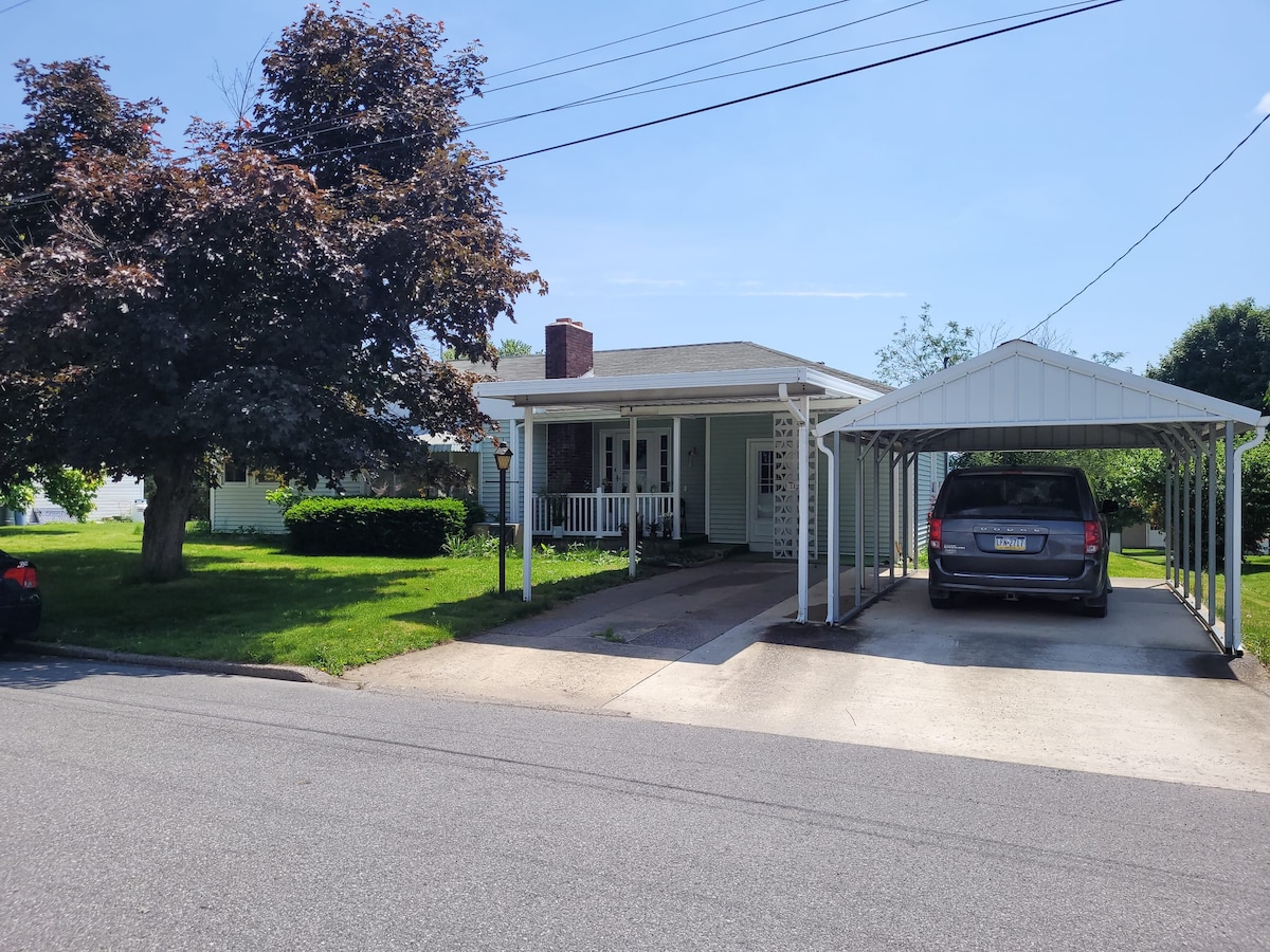 A single-story home featuring a front porch with white railings, surrounded by grassy landscaping and mature trees. An attached carport provides covered parking. The driveway leads from the street to the entrance, ensuring easy access from the road.