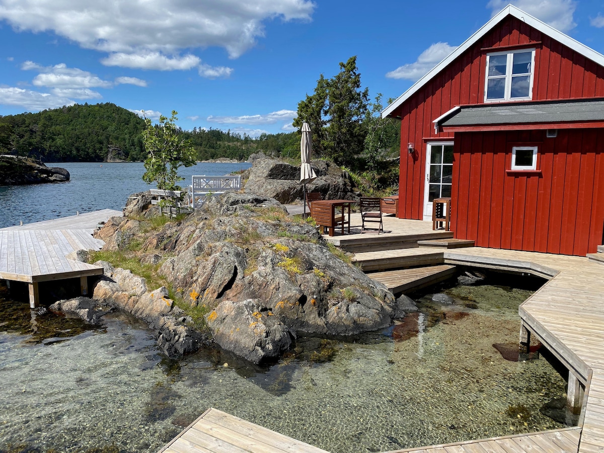 A red wooden building is positioned by the water’s edge, surrounded by lush greenery and rocky outcrops. Wooden walkways extend towards the water, providing access. Sunlight reflects off the clear water, creating a serene environment suitable for relaxation.