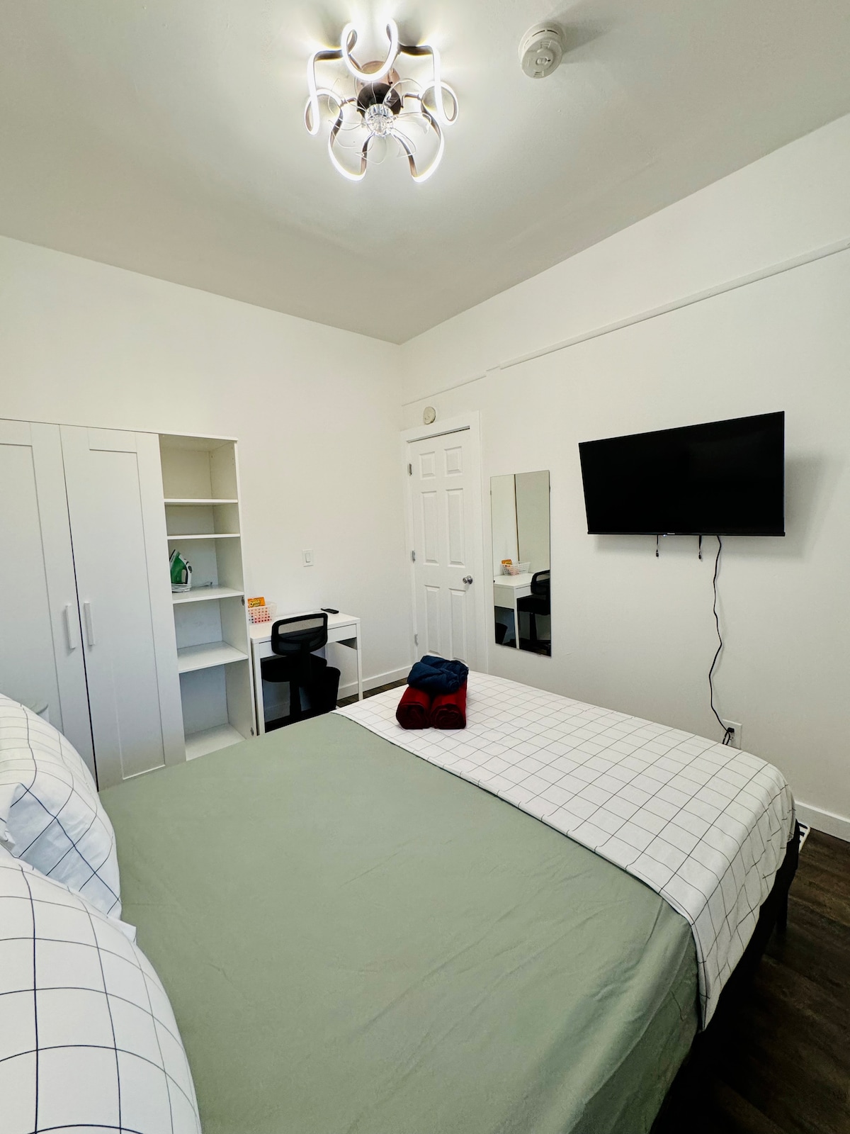 A well-furnished private bedroom is showcased, featuring a spacious bed with a light green coverlet. A wall-mounted TV is positioned opposite the bed. A desk and chair are situated by the closet, complemented by a modern light fixture overhead.