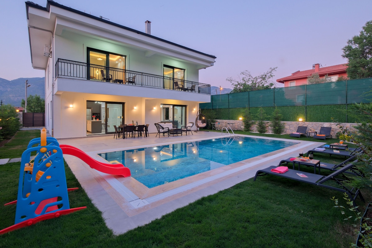 A modern two-story villa features large glass doors opening to a private pool area. A colorful slide and sun loungers are set beside the pool, which is surrounded by well-maintained grass. The inviting outdoor space benefits from gentle lighting as dusk approaches.