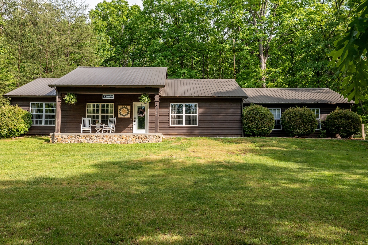The exterior of the Thompson House is shown, featuring a welcoming porch with rocking chairs and a neatly landscaped front yard. The setting is surrounded by trees, showcasing lush greenery and a well-maintained lawn that leads to the entrance.