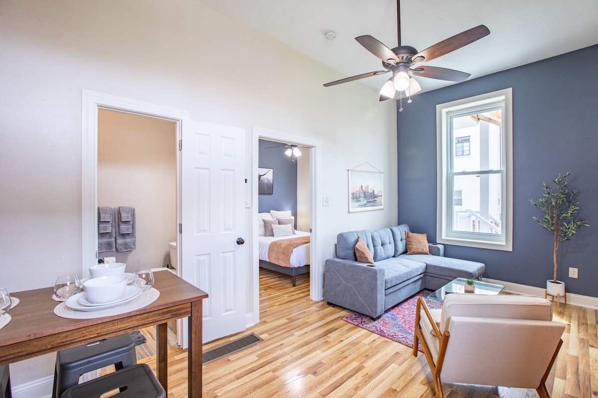 A living space featuring a gray sectional sofa and a light wood table set for dining. An area rug adds color to the hardwood floor. A window allows for natural light, and a doorway leads to a bedroom area with a spacious bed visible.