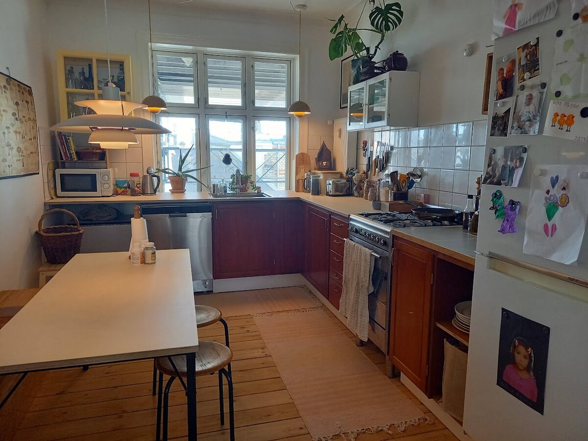 A bright kitchen area features wooden cabinets and a spacious table with metal chairs. Natural light floods through large windows, illuminating various kitchen appliances and decor. Artwork is displayed on the walls, adding a personal touch to the inviting space.