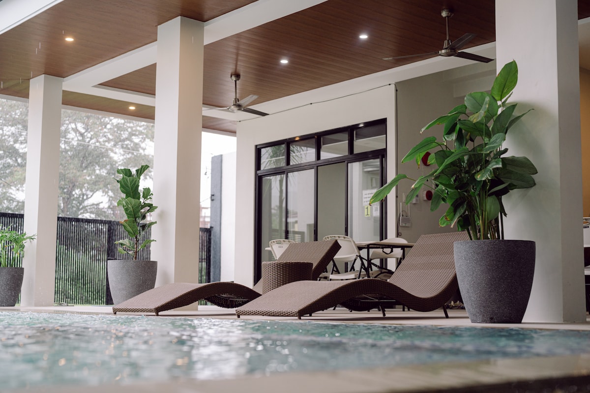 A covered patio area is visible, featuring two lounge chairs positioned beside a tranquil pool. Potted plants provide greenery, and a dining table can be seen through large glass doors, allowing for seamless indoor and outdoor living.