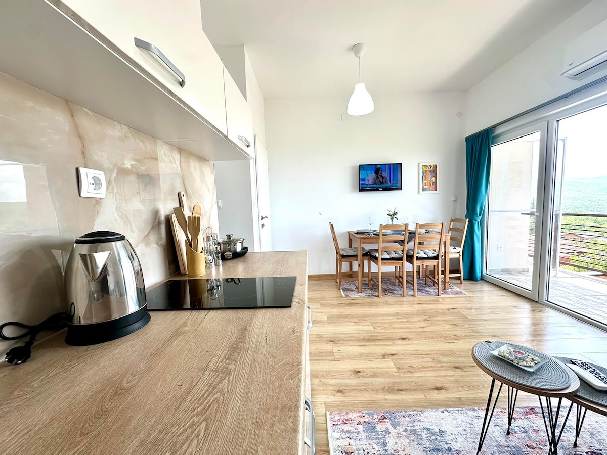 A modern kitchen area featuring sleek cabinetry and a stovetop, with a kettle and utensils visible. Adjacent, a dining space is furnished with a wooden table and matching chairs, while a large window provides natural light and scenic views of the surroundings.
