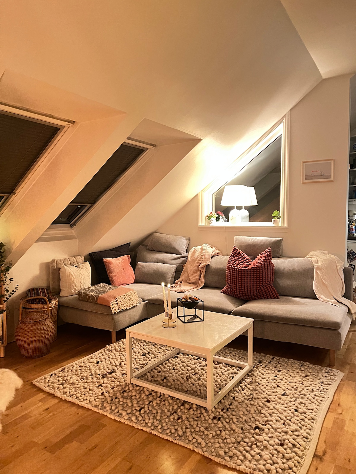 A comfortable living area features a sectional sofa adorned with decorative pillows and throws. A white coffee table sits on a textured rug, accompanied by a small basket. Natural light enters through angled windows, and a lamp provides additional illumination.