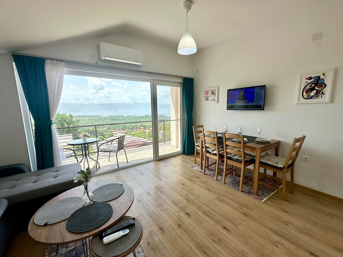 A bright living space features large glass doors that open to a scenic view. Light wood flooring complements a dining area with a modern table and chairs. A cozy sofa is positioned nearby, and a wall-mounted television is visible, along with decorative art pieces.