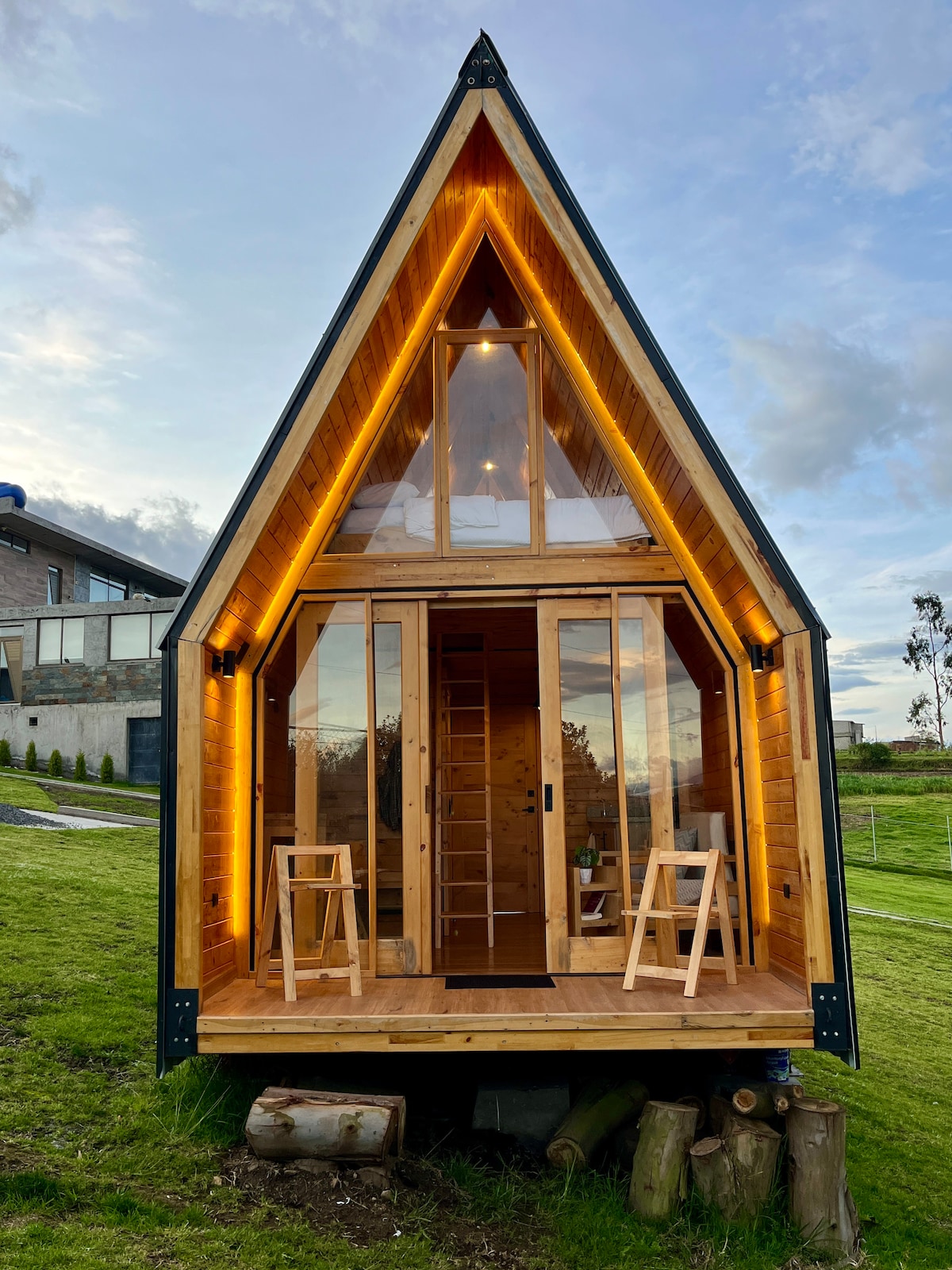 The exterior of a triangular shaped tiny home is showcased, featuring wooden panels and large front windows that allow ample natural light. Two wooden stools are positioned on a small porch under warm, ambient lighting, providing a welcoming entrance amid an expansive green landscape.