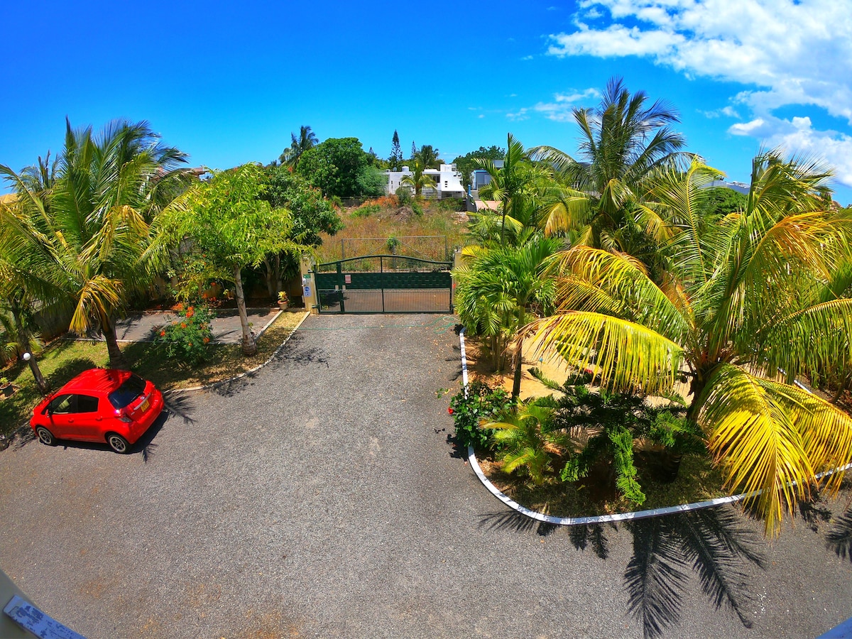 A spacious garden area is surrounded by lush palm trees and greenery, providing a serene environment. A red car is parked on the gravel driveway, leading to a secure entrance gate. The bright blue sky creates a vibrant contrast with the landscape.