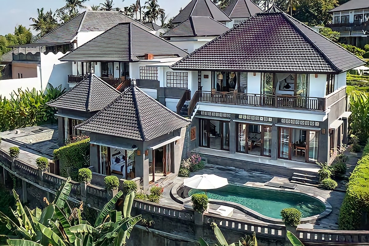This aerial view showcases the spacious villa with a private pool set amid lush greenery. The modern architecture features multiple levels, large windows, and open outdoor spaces. A sun lounger beside the pool invites relaxation within the serene landscape.