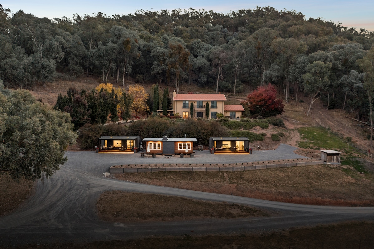 Aerial view of the accommodation property is displayed, featuring multiple cabins set against a backdrop of lush greenery. The main house is positioned prominently, with autumn foliage visible in the surrounding landscape. A gravel path leads to the cabins, emphasizing the spacious outdoor setting.