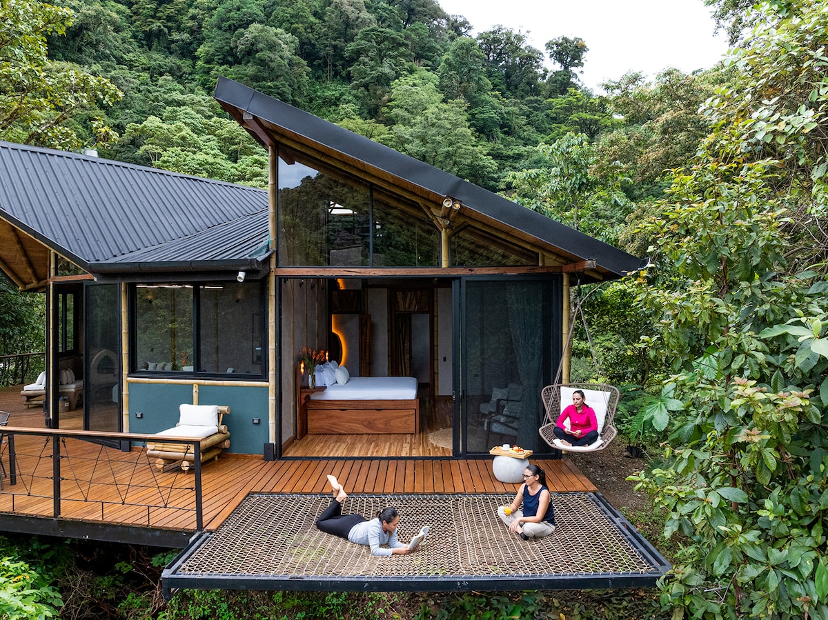 A spacious wooden deck extends from a modern cabin, with large glass doors providing an open view of the serene jungle surroundings. A comfortable hammock hangs nearby, while two individuals relax on the deck, immersed in nature. The cabin's design combines natural elements and contemporary architecture.