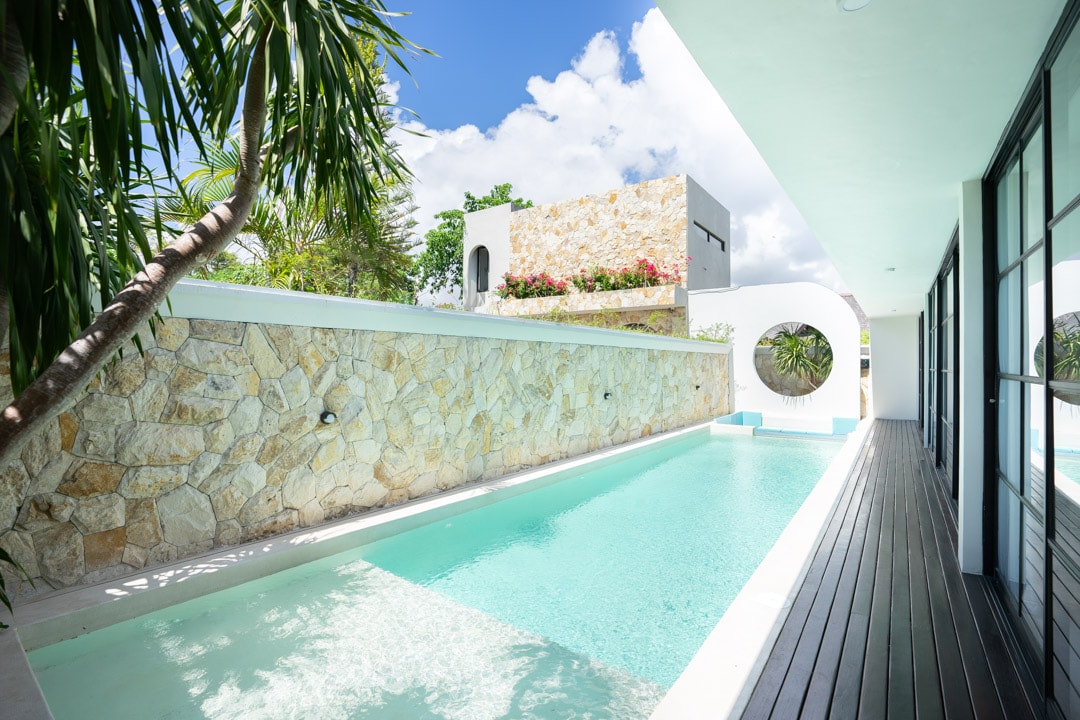 A striking outdoor area features a clear swimming pool bordered by natural stone walls, surrounded by tropical greenery. Sunlight reflects off the water, creating a serene atmosphere. The modern architecture includes wide glass doors, allowing views of the pool from the adjoining indoor space.