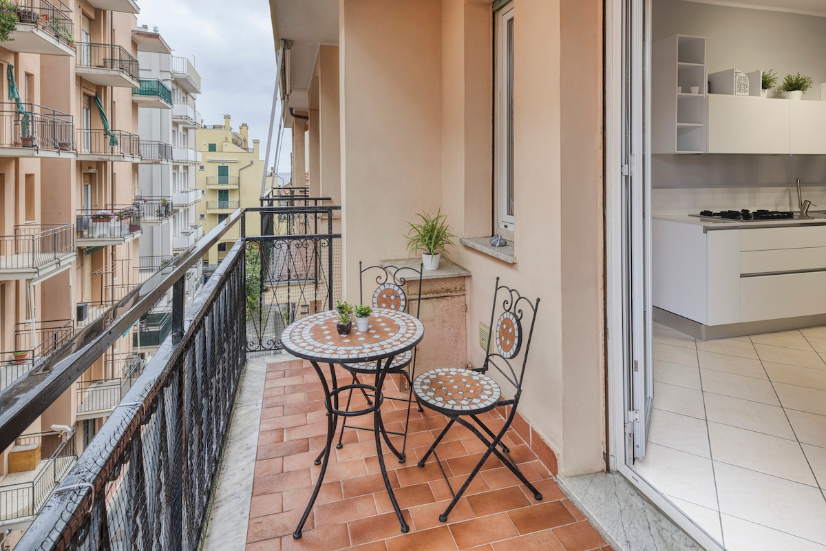 A small terrace features a round table and two metal chairs, positioned on terracotta tiles. Nearby, a spacious kitchen is visible through glass sliding doors, enhancing the connection between indoor and outdoor spaces.
