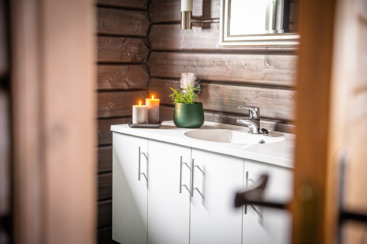 A clean and modern bathroom features a white vanity with multiple drawers and a white sink. Soft candlelight from three candles enhances the serene atmosphere. A green plant adds a touch of nature, while a large mirror reflects the space.