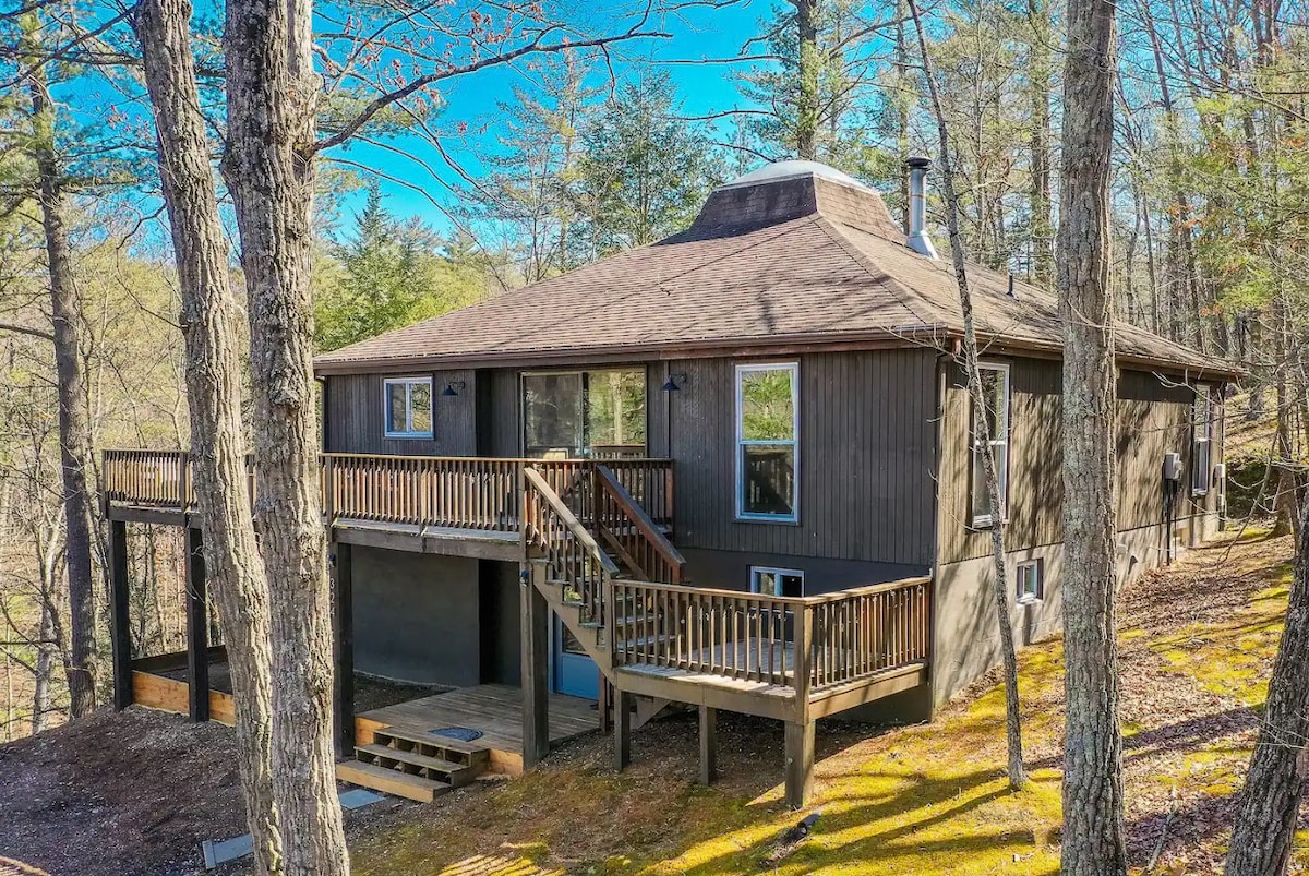 The cabin features a wood exterior set amongst tall trees, showcasing a wraparound deck with multiple entry points. Large windows allow natural light to fill the interior, while a sloped landscape enhances its private and serene setting.