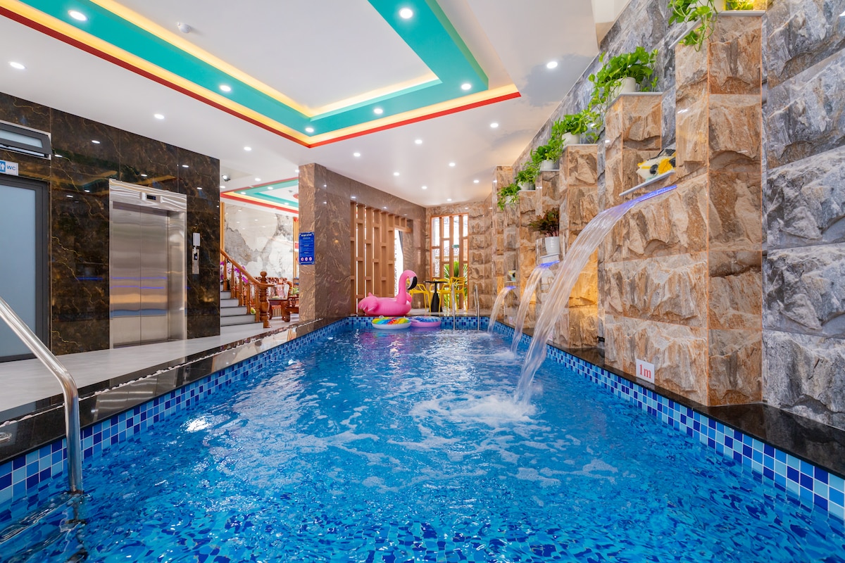 An indoor pool features vibrant blue tiles and rock formations along the water's edge. Water cascades gently from the rocks, creating a soothing atmosphere. A pink inflatable float rests on the surface, and seating is visible in the background.