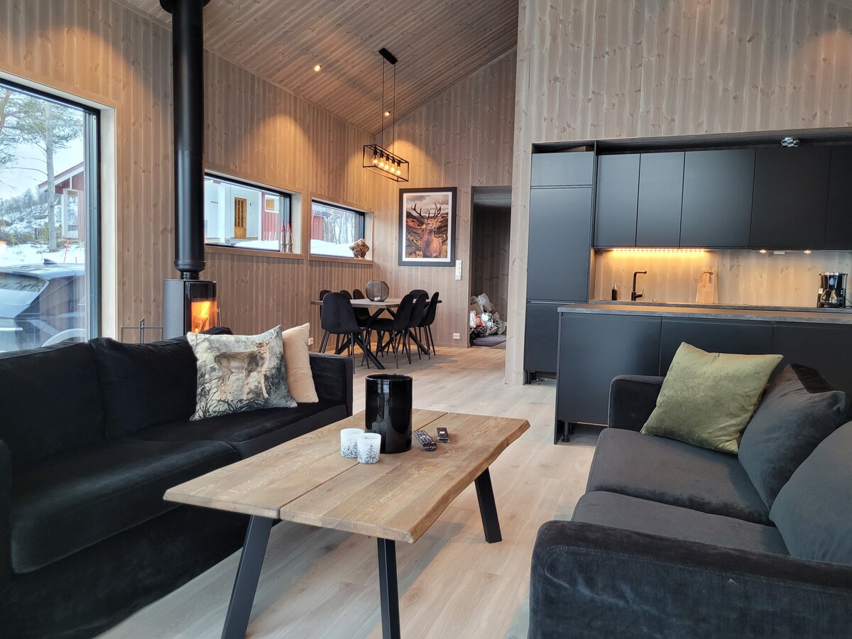 An open living area features two dark sofas and a wooden coffee table with decorative items. A dining space is visible in the background with a round table set for six. The modern kitchen is sleek with dark cabinetry and warm lighting, enhancing the space.