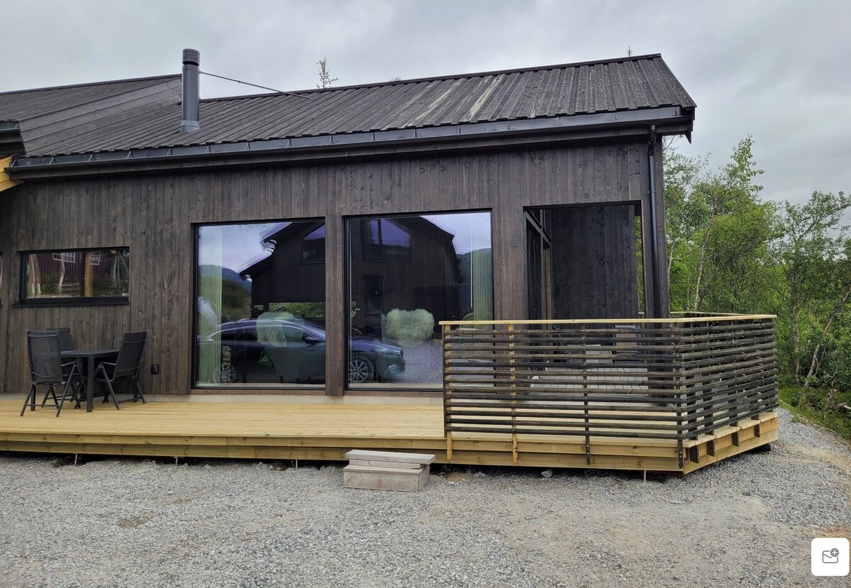 The exterior of the cabin features a wooden deck with a modern railing, leading to large glass windows that offer views of the surrounding landscape. A small seating area with two chairs and a table is visible on the deck, creating an outdoor gathering space.
