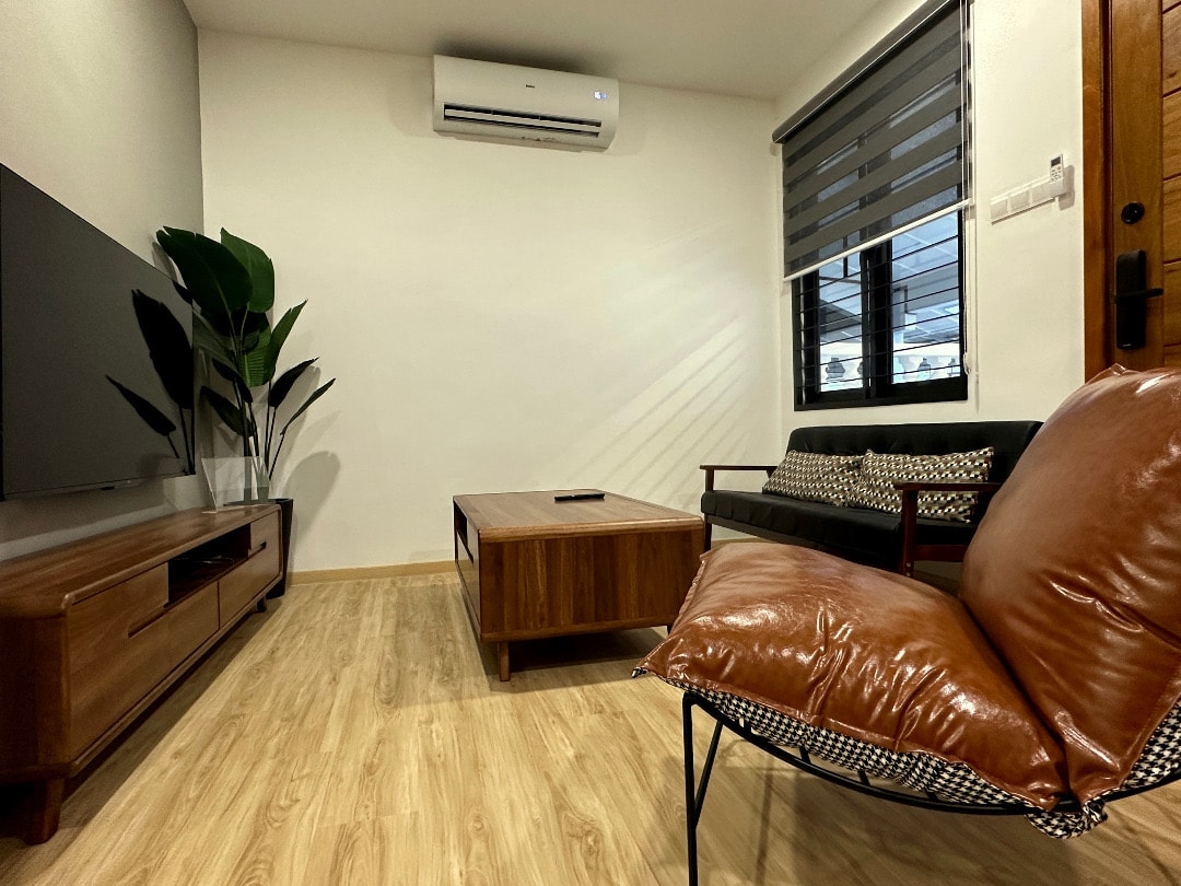The living room features a wooden TV cabinet with a flat-screen television mounted on the wall. A comfortable black sofa sits across from a rectangular coffee table. A stylish armchair is positioned nearby, while a tall indoor plant adds a touch of greenery to the space.