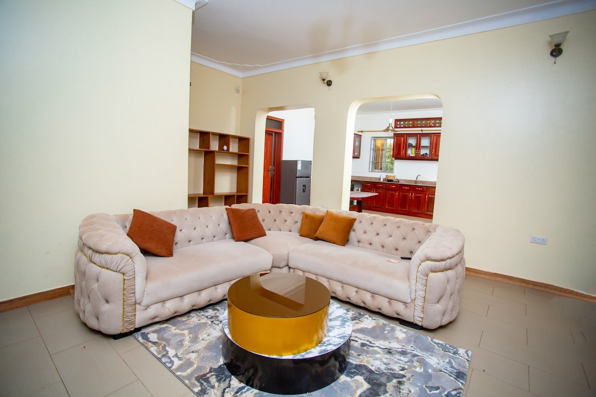 A spacious living area features a plush, curved sofa in neutral tones, accented by decorative pillows. A circular coffee table with a reflective surface sits on a patterned rug, creating a cozy focal point. The open layout provides a view of the adjoining kitchen space.
