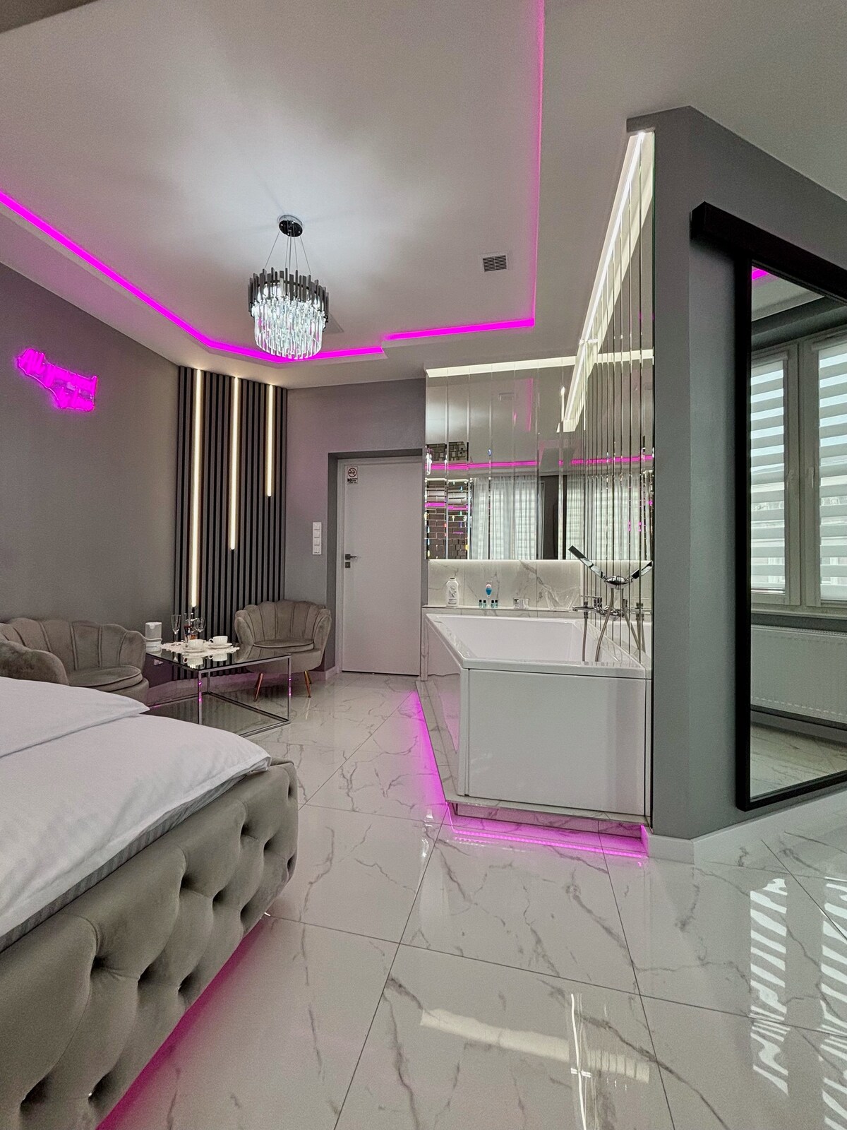 A modern interior showcases sleek white cabinetry and polished marble flooring. Pink lighting highlights the ceiling and mirrors, enhancing the contemporary design. Two stylish armchairs are positioned beside a small table, creating a cozy seating area near the window, which provides natural light.