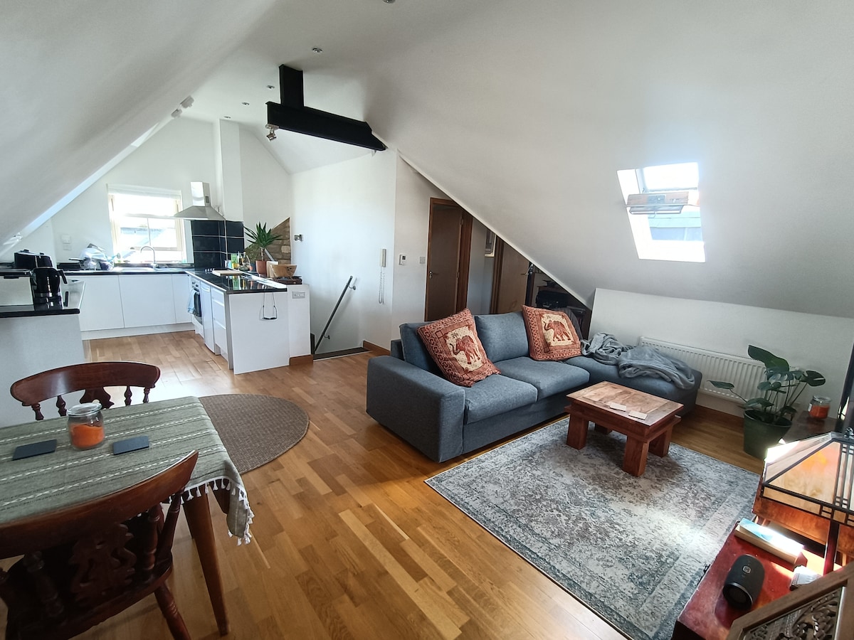 A well-lit living area features a sloping ceiling and hardwood floors. A grey sofa is positioned next to a wooden coffee table, with decorative pillows adding a touch of comfort. The open kitchen is visible in the background, showcasing modern appliances and a clean design.