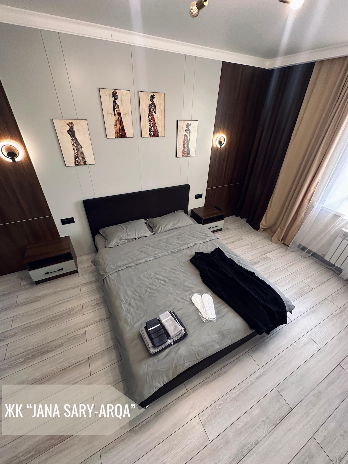 A cozy sleeping area is displayed, featuring a spacious bed with gray linens and two nightstands. Decorative wall art above the bed complements the warm wood accents. Soft lighting from wall sconces and natural light enters through a curtain-covered window.