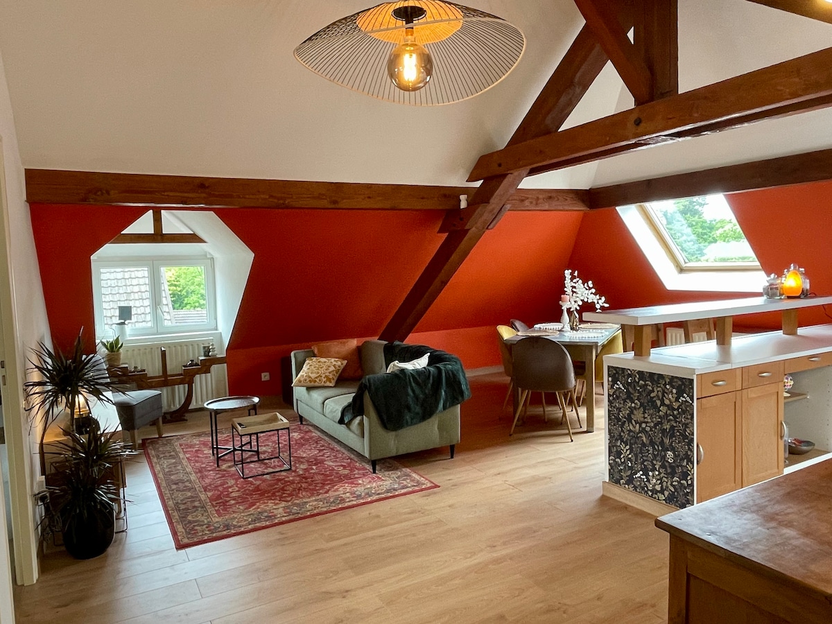 A warm living area is characterized by exposed wooden beams and a sloped ceiling. A comfy sofa is complemented by a small coffee table and decorative plants. A vibrant red accent wall contrasts against the neutral decor, while natural light floods in through a skylight.