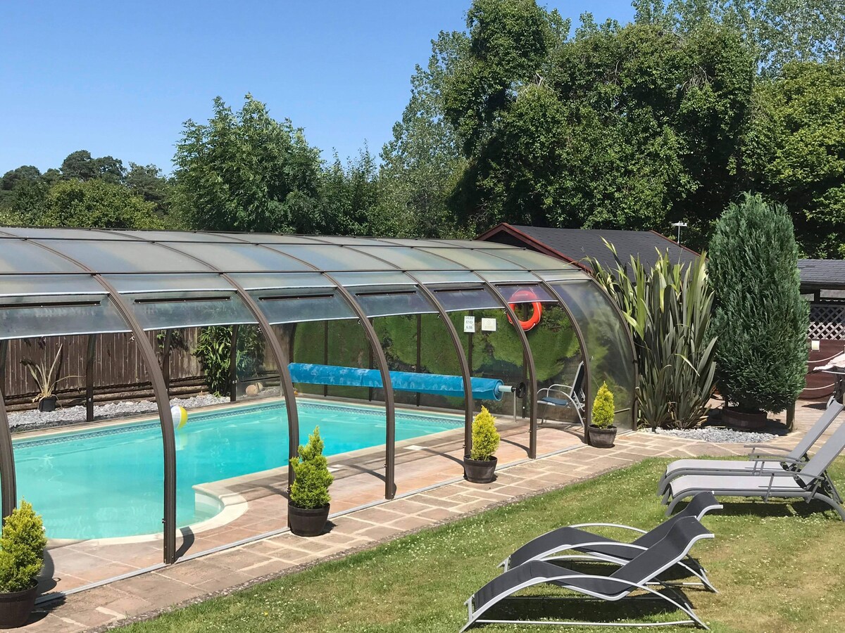 A covered swimming pool is surrounded by neatly trimmed grass, with lounge chairs positioned nearby. Lush greenery frames the area, and a small decorative shrub is planted in pots around the poolside. The protective enclosure allows for year-round enjoyment.