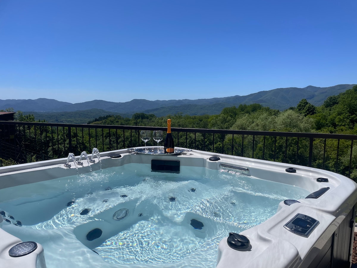 A spacious jacuzzi is positioned on a terrace, providing a panoramic view of green hills and mountains in the distance. Clear blue skies reflect in the water, while sparkling glasses and a bottle of champagne rest on the edge, highlighting a relaxing atmosphere.