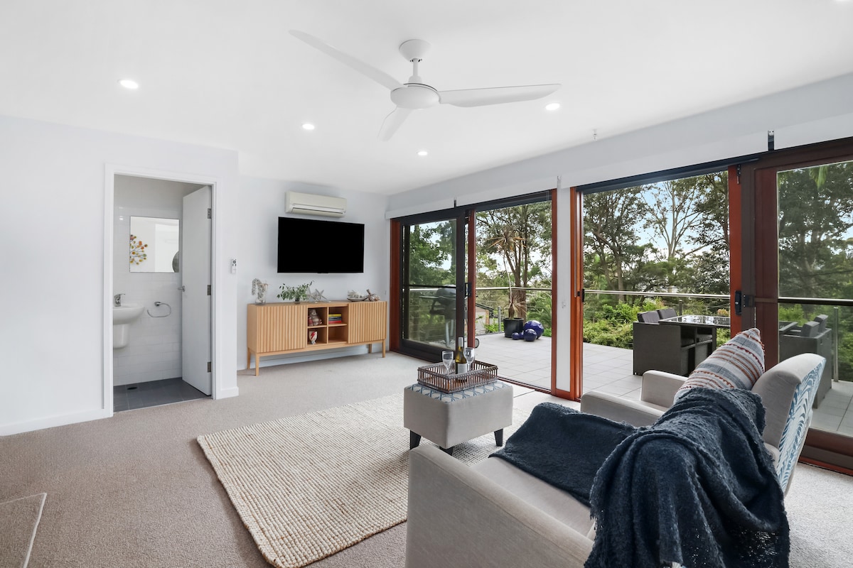 An open living area features large glass doors leading to a deck, with natural light illuminating the space. A plush sofa is accompanied by a textured throw, and a wall-mounted TV is visible. A separate doorway leads to a private bathroom area.