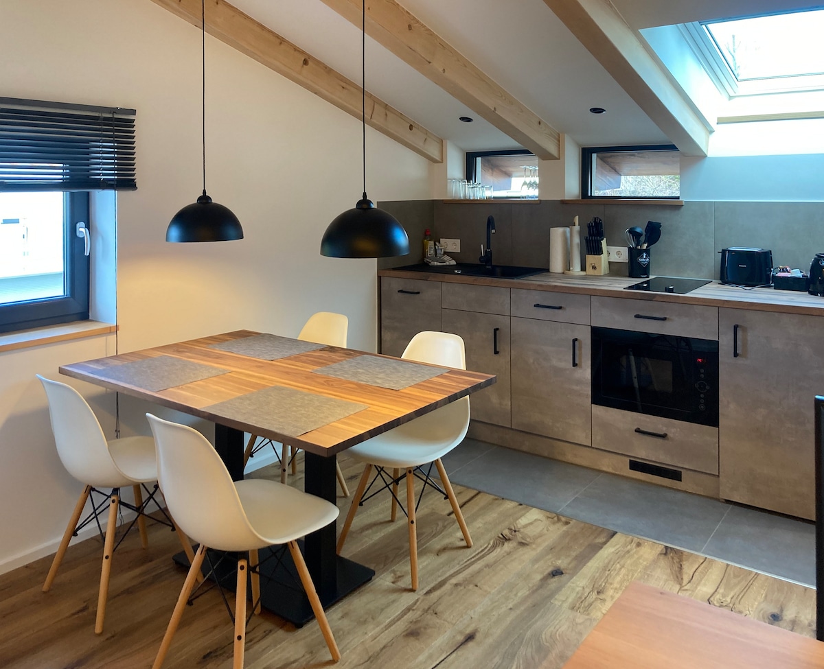 The kitchen and dining area features a wooden table with four white chairs, set under two black pendant lights. A modern kitchen with grey cabinets includes a built-in oven and coffee maker, with sunlight filtering through the skylights and windows.