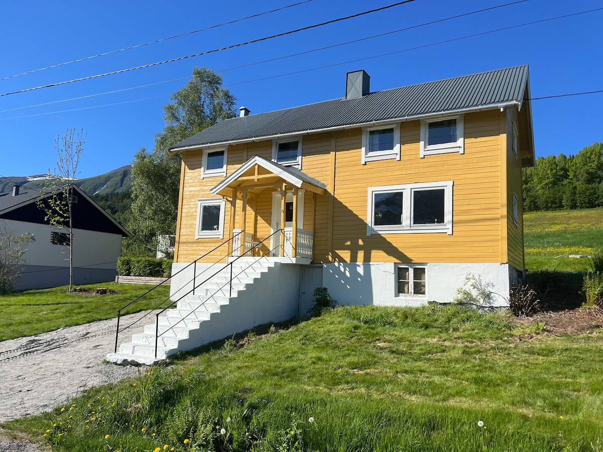 A charming two-story house in a bright yellow exterior is set against a clear blue sky. The entrance is accessible by a wide staircase with a covered porch. Lush green grass surrounds the property, complemented by a scenic hillside in the background.