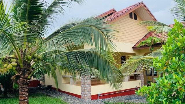 The exterior of the apartment is showcased, featuring a well-maintained garden with lush palm trees. The building's facade is designed with a combination of beige and red tones, and a patio is visible, providing a welcoming entrance to the home.