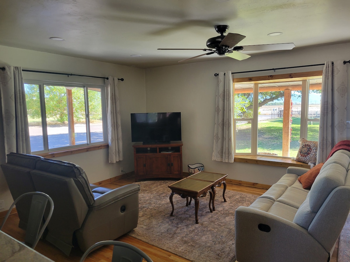 A comfortable living area is enclosed by large windows, allowing natural light to fill the space. A light-colored sofa and armchairs are arranged around a wooden coffee table. A flat-screen television is mounted on the wall, enhancing the room's functionality.