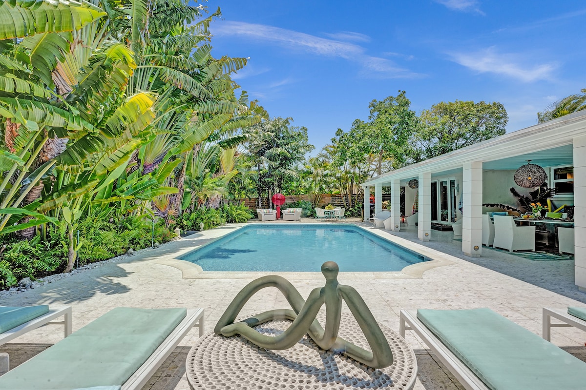 A spacious outdoor area is presented featuring a heated pool surrounded by tropical greenery. Sun loungers are positioned around the pool deck, enhancing the inviting atmosphere. The backyard is complemented by an open seating area and a modern artistic sculpture, promoting relaxation and leisure.