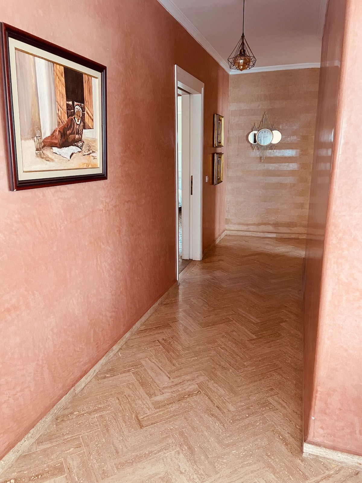 A hallway is highlighted, featuring warm peach-colored walls and a polished herringbone wooden floor. Decorative elements include framed artwork, vintage-style mirrors, and ambient lighting provided by a pendant fixture. The open door at the end leads to another area of the apartment.