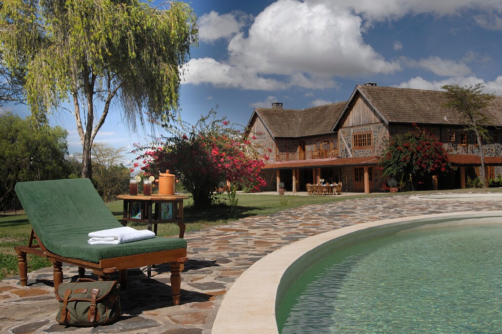 A lounge chair is positioned beside a natural stone pool, accompanied by a table displaying refreshments. In the background, a wooden structure with a sloped roof is partially surrounded by vibrant greenery and flowering plants. Fluffy clouds add texture to the sky above.