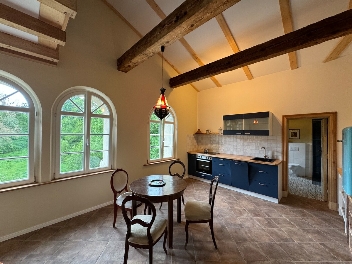 A spacious dining area features a round wooden table surrounded by four chairs. Natural light pours in through large arched windows, illuminating the white and blue cabinetry in the adjoining kitchen. Exposed wooden beams along the ceiling add architectural interest to the room.