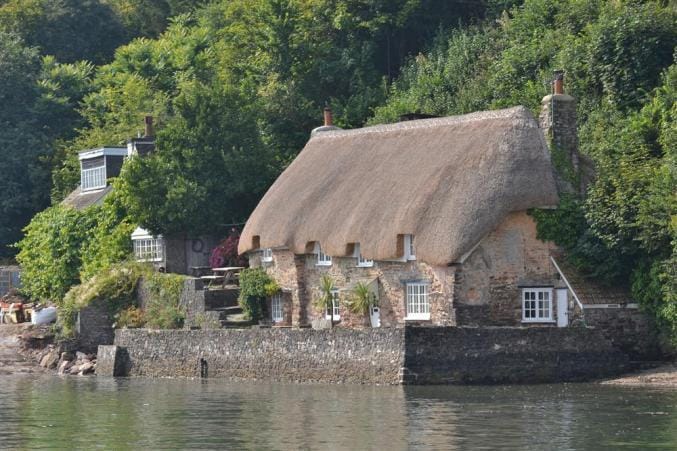A charming thatched cottage is nestled alongside the river Dart, featuring lush greenery and stone walls. The building's thatched roof complements its rustic design, while windows reflect the surrounding scenery. A pathway leads to the rear, enhancing the serene riverside setting.