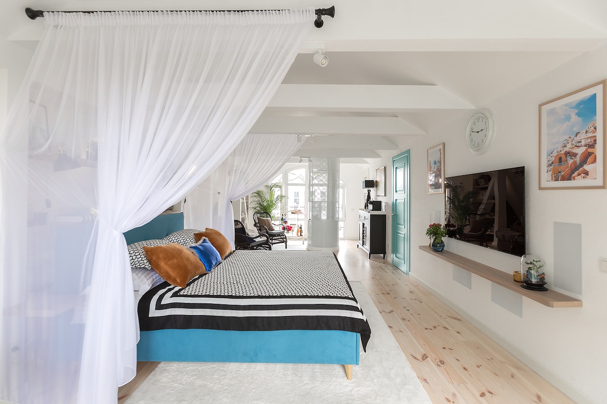 A spacious bedroom features a large bed with decorative pillows and a contrasting bedspread. Sheer white curtains frame the space, allowing light to filter through. A television is mounted on the wall, while a small plant and decorative pieces sit on the shelf below it.