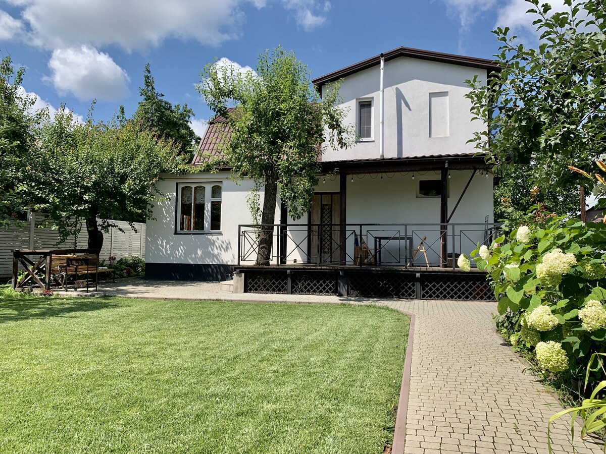 A two-story house features a well-maintained garden with a manicured lawn and blooming hydrangeas. A wooden deck is accessible from the main entrance, flanked by seating areas under the shade of trees. Natural light brightens the exterior, creating a welcoming ambiance.