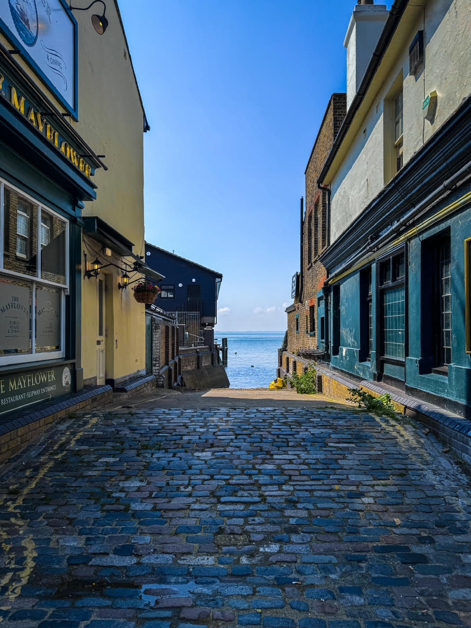 A narrow cobblestone alleyway is flanked by colorful buildings, leading towards the sea. The bright blue sky is visible above, while the shimmering water can be seen at the end of the pathway, inviting exploration.