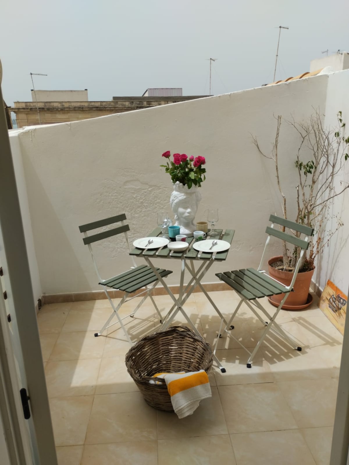 A small outdoor terrace is furnished with a glass table and two green folding chairs. A white vase filled with pink roses sits at the center of the table, complemented by tableware. A decorative basket is placed on the floor, adding to the inviting atmosphere.
