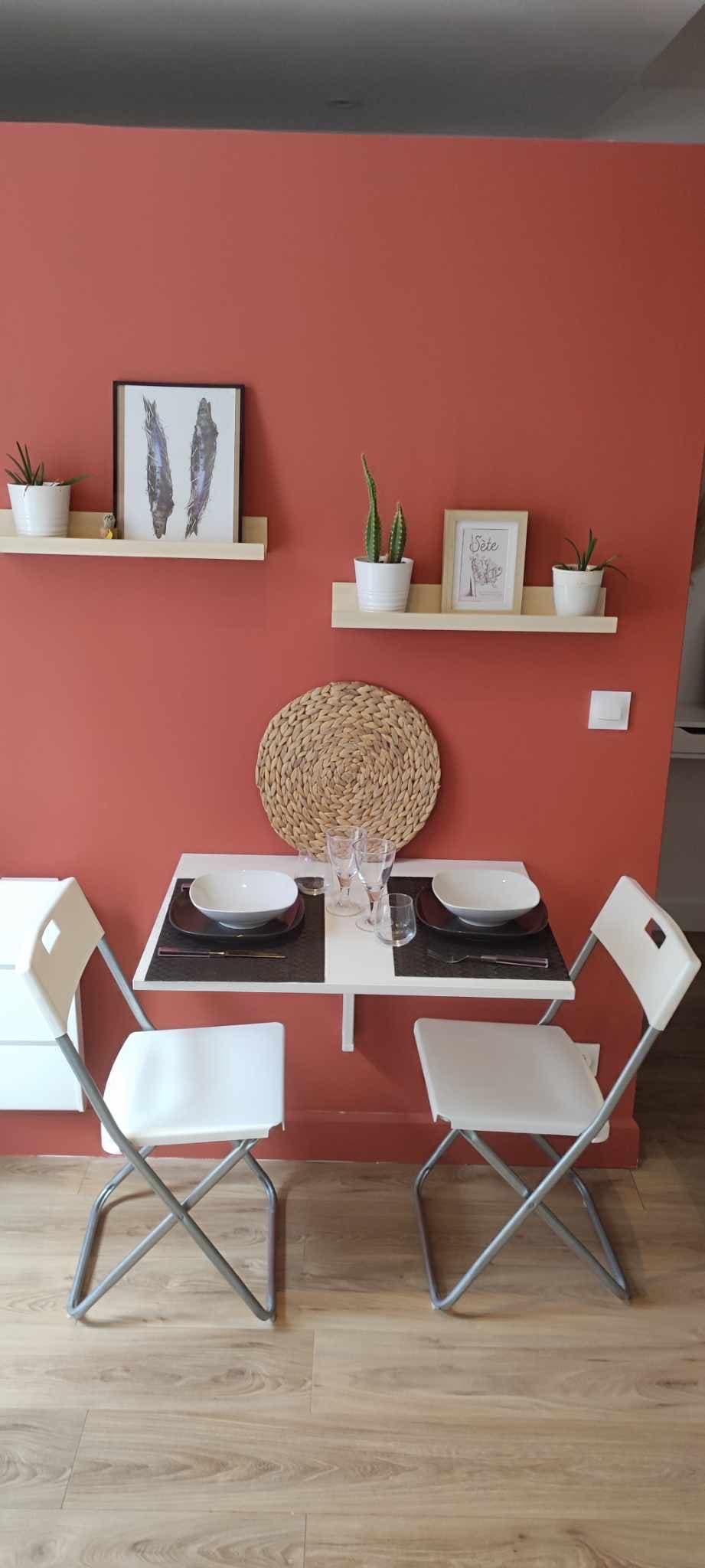 A small dining area is present with a foldable table set against an orange wall. Two white chairs are positioned at the table, which is set with plates and glassware. Green plants are displayed on wall shelves above the table, adding a touch of greenery.