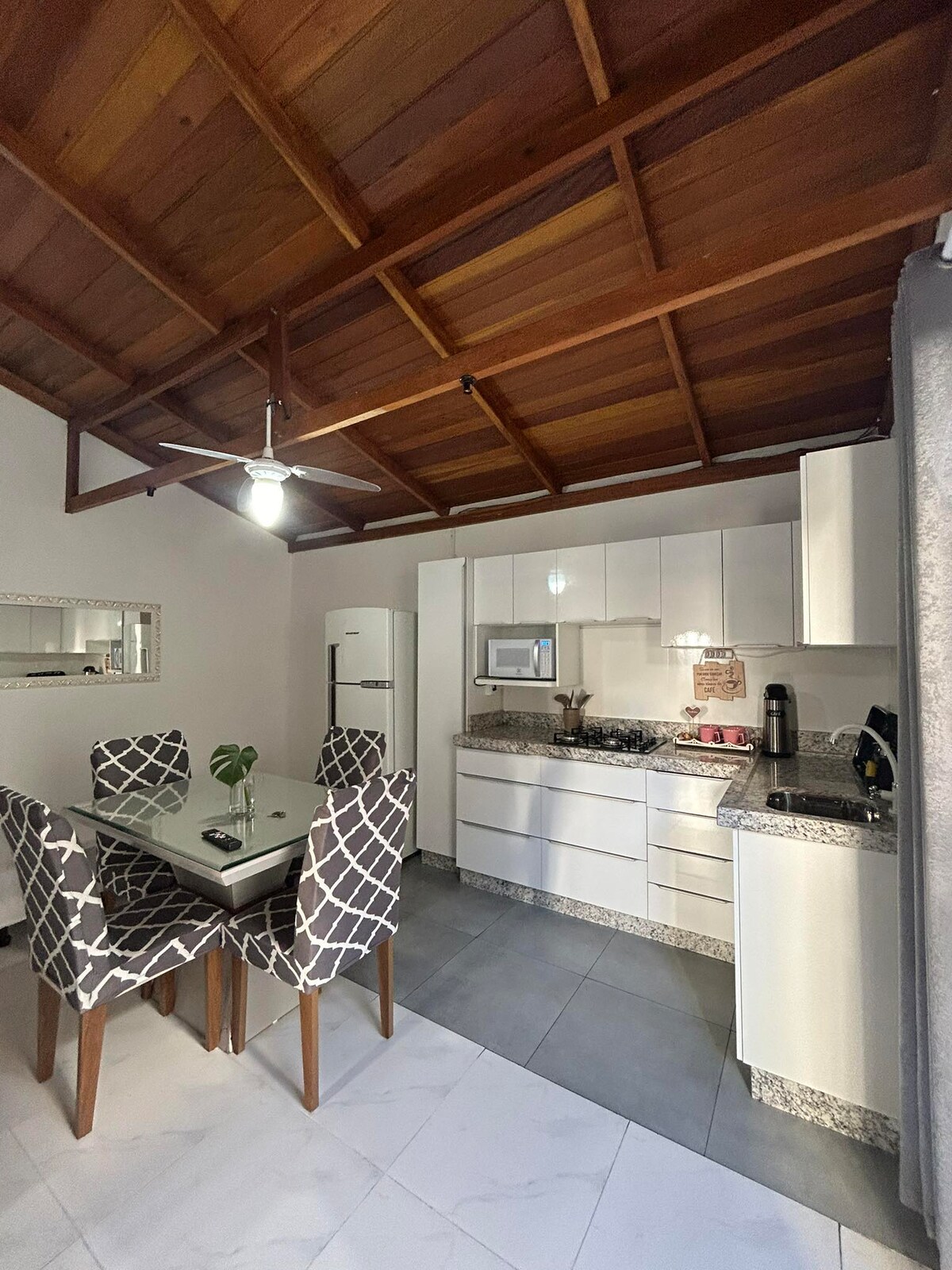 A modern kitchen and dining area feature a glass dining table surrounded by four upholstered chairs. The kitchen is equipped with sleek white cabinets, a microwave, and a stove. Exposed wooden beams add a touch of warmth to the space, enhanced by natural light from a window.
